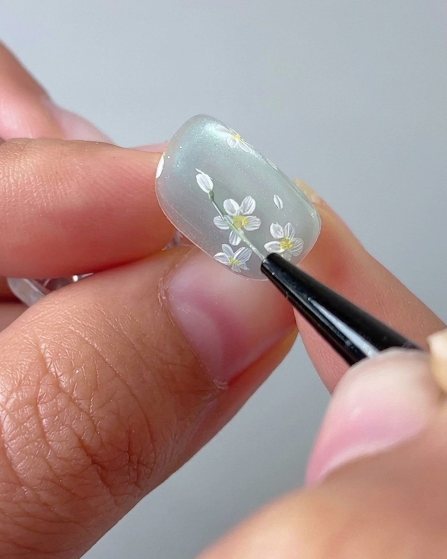Eine Nahaufnahme eines Nagelpinsels, der einen dünnen grünen Stängel auf einer Cat-Eye-Gelbasis zeichnet.
