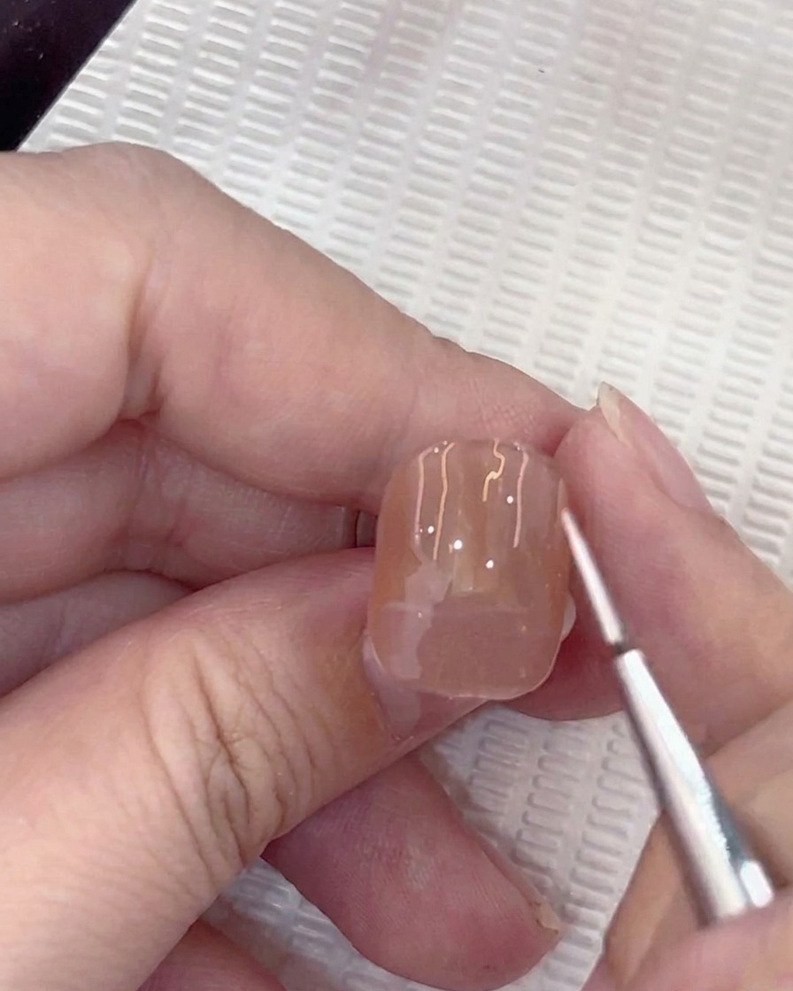 Eine Nahaufnahme eines Linierpinsels, der einen kleinen weißen Führungspunkt in die Mitte eines Nude-Gelnagels setzt.