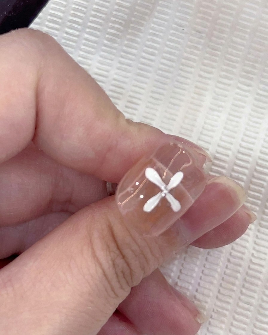 Nahaufnahme eines Nagels mit fünf weißen Blütenblättern in kreisförmigem Muster, während ein Pinsel ein sechstes Blatt hinzufügt.
