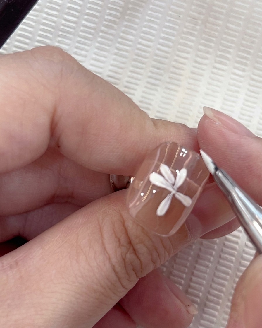Nahaufnahme eines weißen Gänseblümchen-Nageldesigns, bei dem ein siebtes Blütenblatt vorsichtig mit einem Rundpinsel platziert wird.