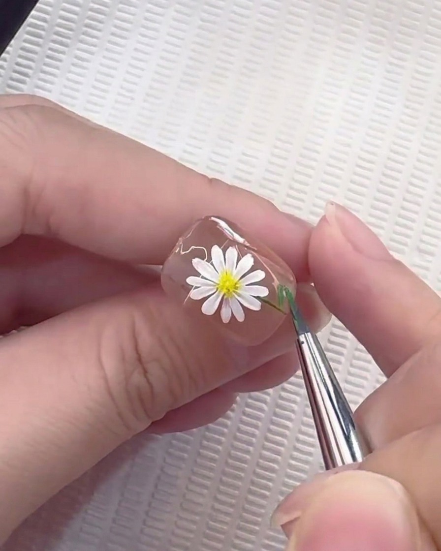 Ein Linierpinsel zieht eine dünne grüne Linie, um einen Stiel auf einem mit Gänseblümchen verzierten Nagel zu bilden.