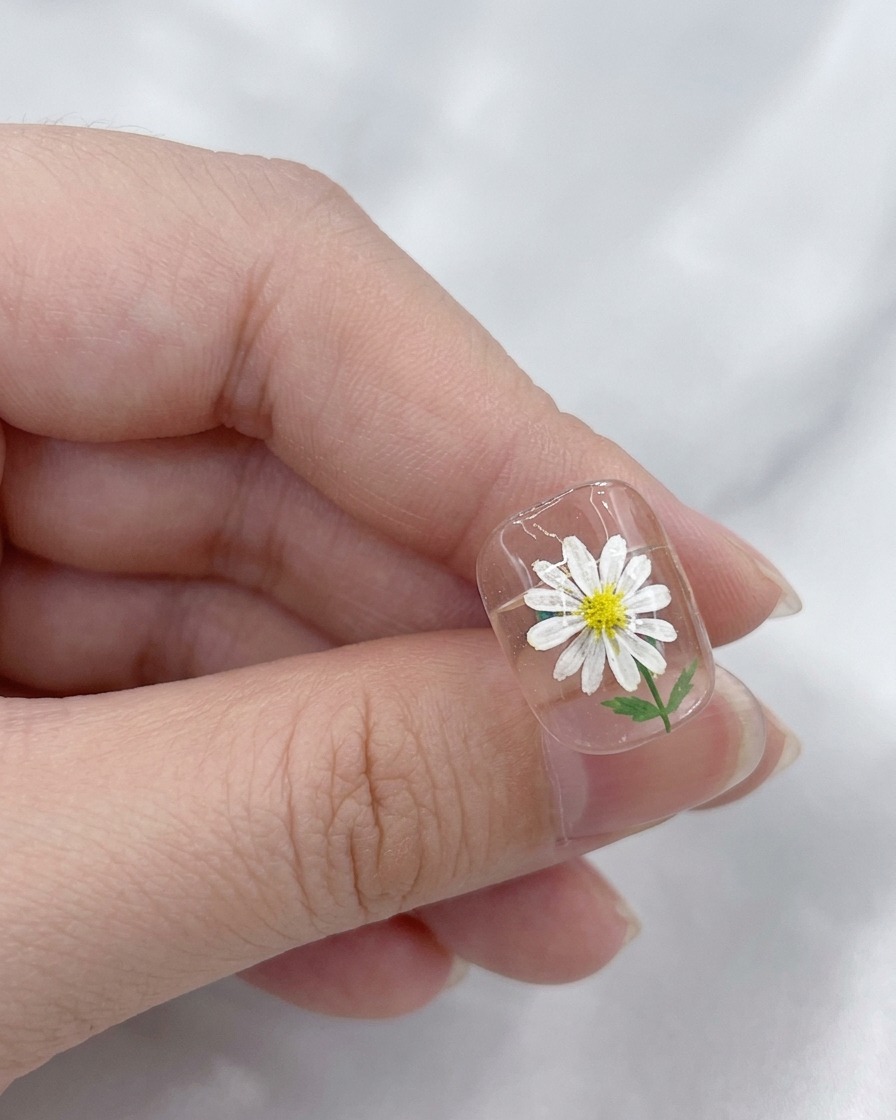 Ein Pinsel trägt einen klaren, glänzenden Top Coat über ein fertiges Gänseblümchen-Nageldesign auf.