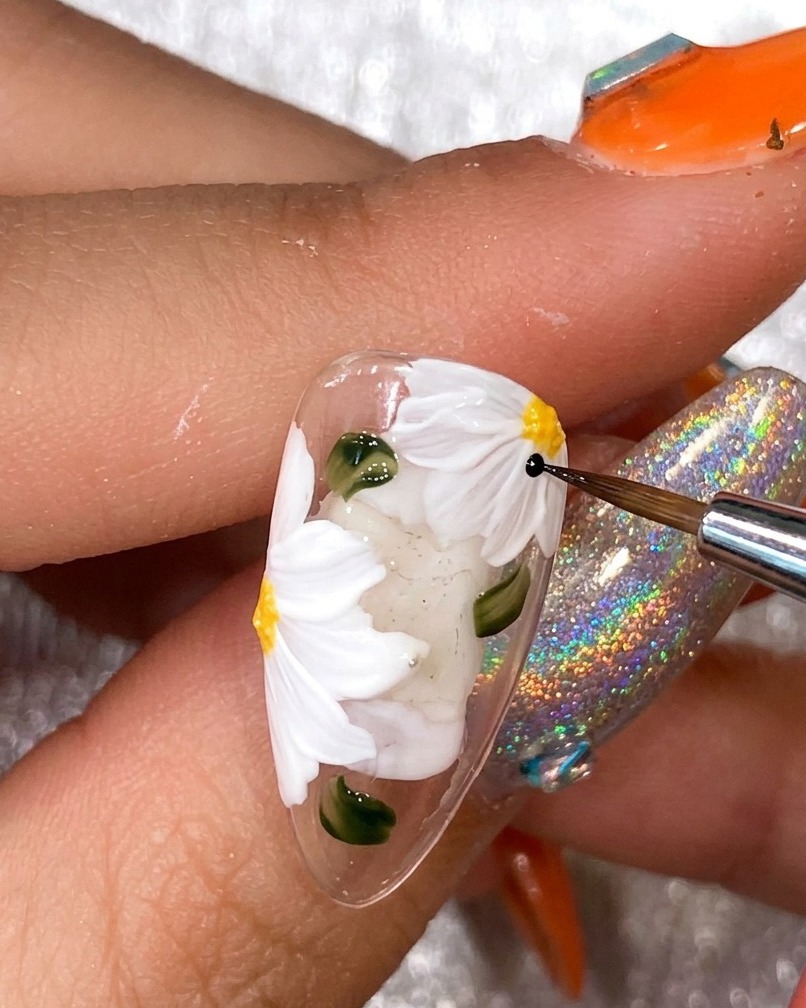 Nahaufnahme der Hand eines Nageldesigners, die mit einem dünnen schwarzen Liner-Pinsel die weißen Gänseblümchen-Blütenblätter auf einem Nagel-Tip umrandet.