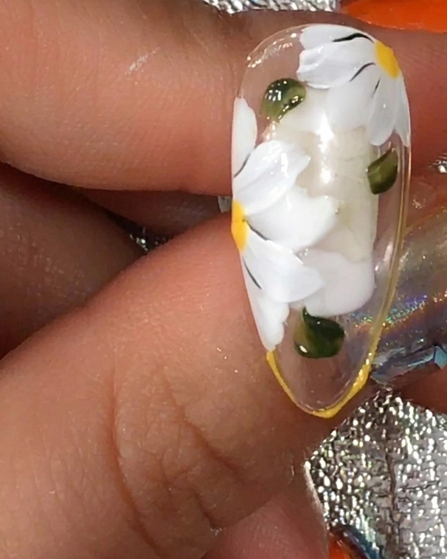 Ein Pinsel trägt eine abschließende Schicht matten Top-Gels über einen Nagel mit weißem Gänseblümchen-Design und grünen Blättern auf.