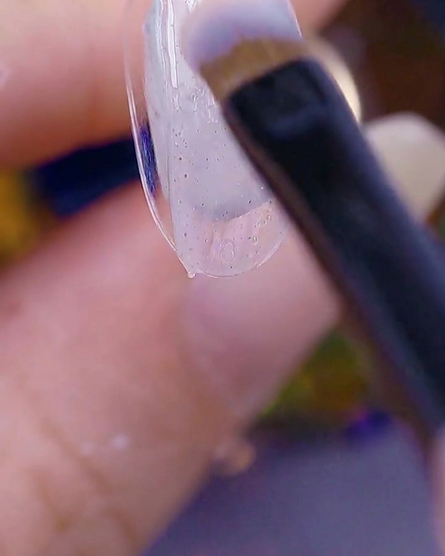 Nahaufnahme einer Bürste, die ein transparentes, texturiertes Sugar-Gel auf die Spitze eines Naturnagels aufträgt.
