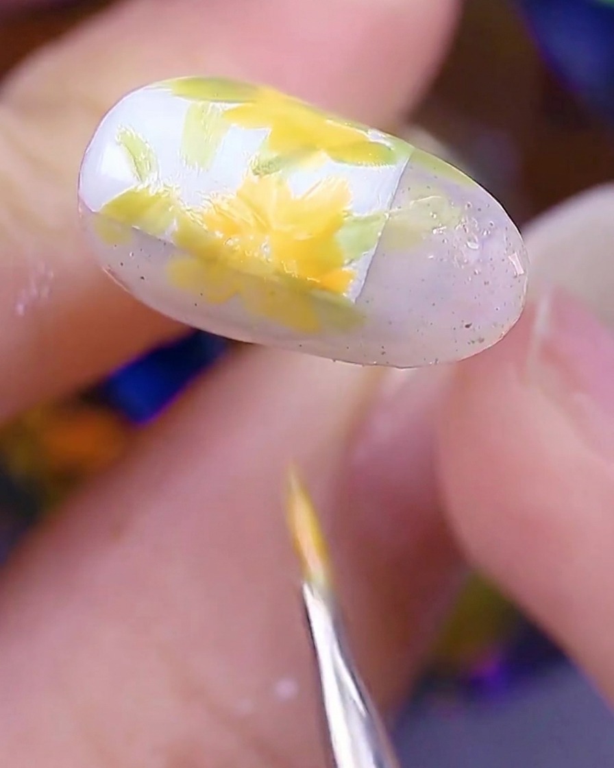 Nahaufnahme des Nagels, die mehrere gelb-orangefarbene Gänseblümchen über den grünen Blattbasen zeigt, wobei der Pinsel die letzten Blütenblätter fertigstellt.
