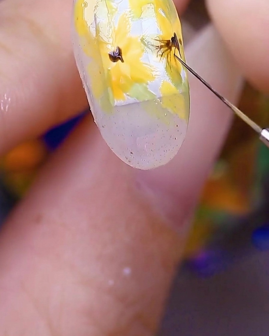 Nahaufnahme der Nailart, die zeigt, wie ein Pinsel feine, dünne dunkelbraune Linien aus der Mitte der gelben Blumen nach außen in die Blütenblätter zieht.