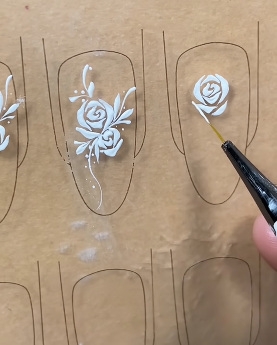 Nahaufnahme eines Liner-Pinsels, der feine weiße Details an den äußeren Blättern des Rosen-Designs auf dem Nageltip hinzufügt.