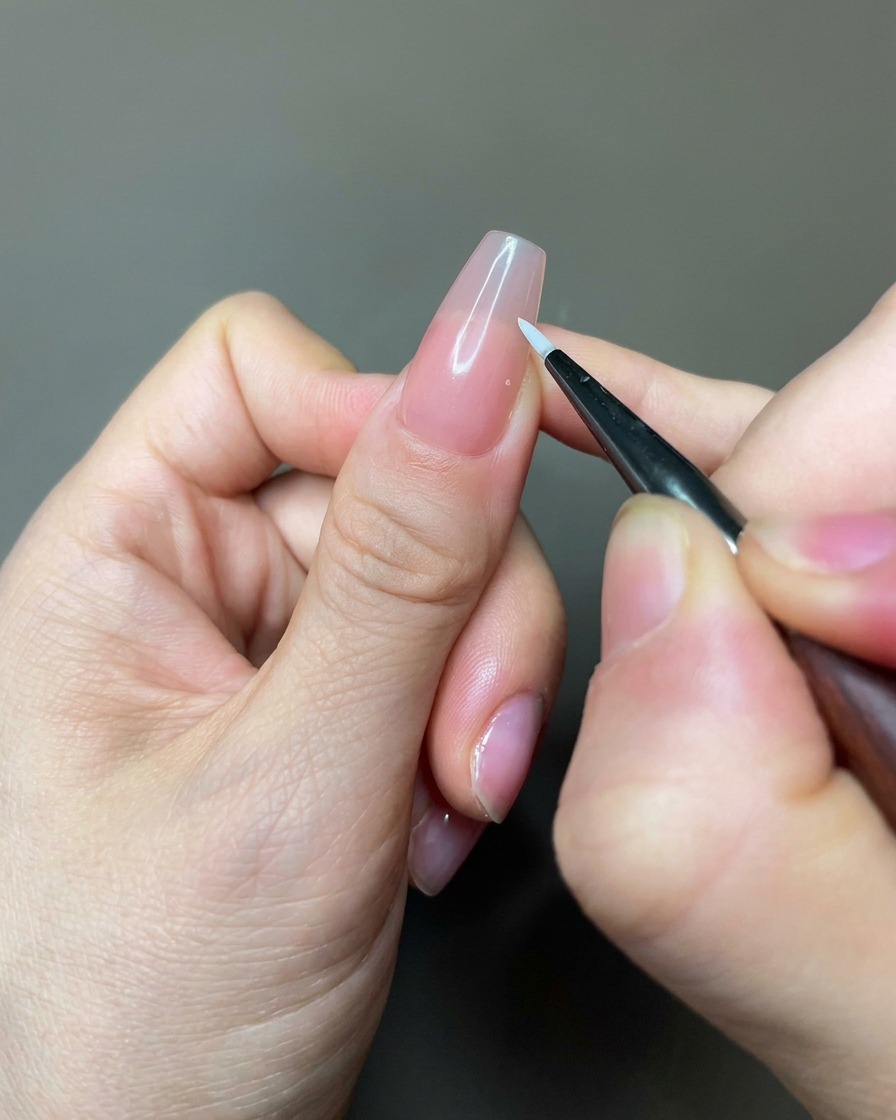 Nahaufnahme eines feinen Liner-Pinsels, der weißes Gel für Blütenblattformen auf einen nudefarbenen Nagel aufträgt.