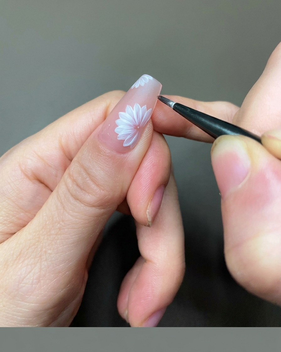 Nahaufnahme einer Naildesignerin, die mit einem feinen Liner-Pinsel goldene Gel-Details in die Mitte eines weißen Gänseblümchens auf einem Nude-Nagel setzt.