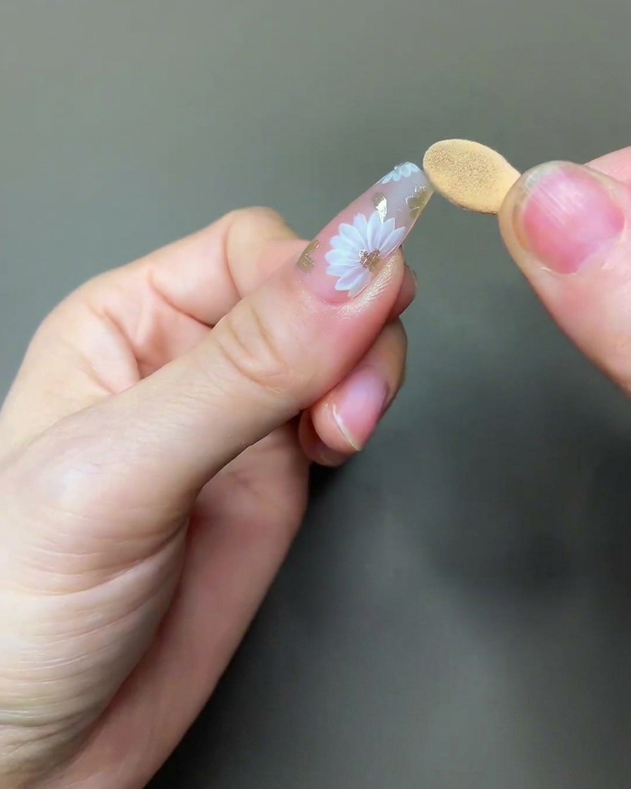 Ein Pinsel trägt einen klaren, glänzenden Top Coat über das fertige Gänseblümchen- und Chrom-Nailart-Design auf.
