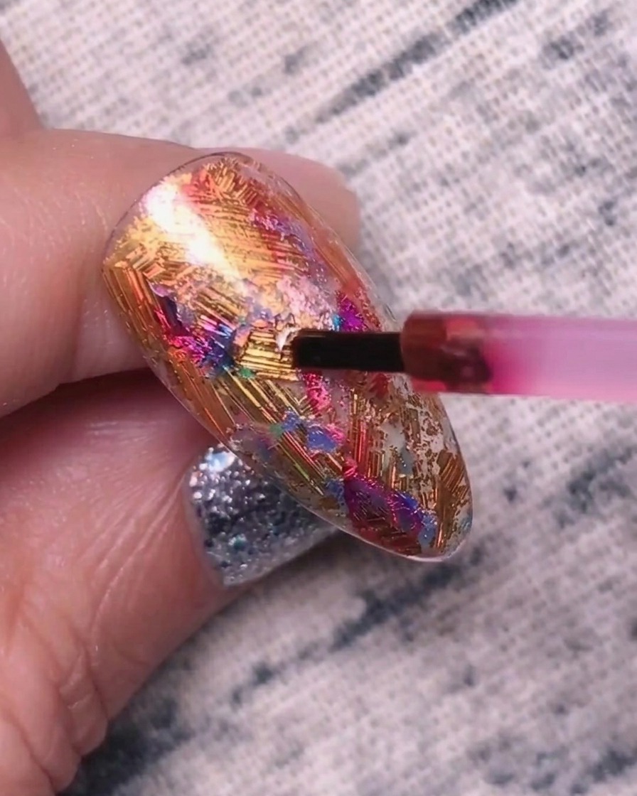 Ein kleiner Pinsel trägt pigmentiertes Rot-Lila-Gel auf eine texturierte, mit Metallfolien bedeckte Nageloberfläche auf.