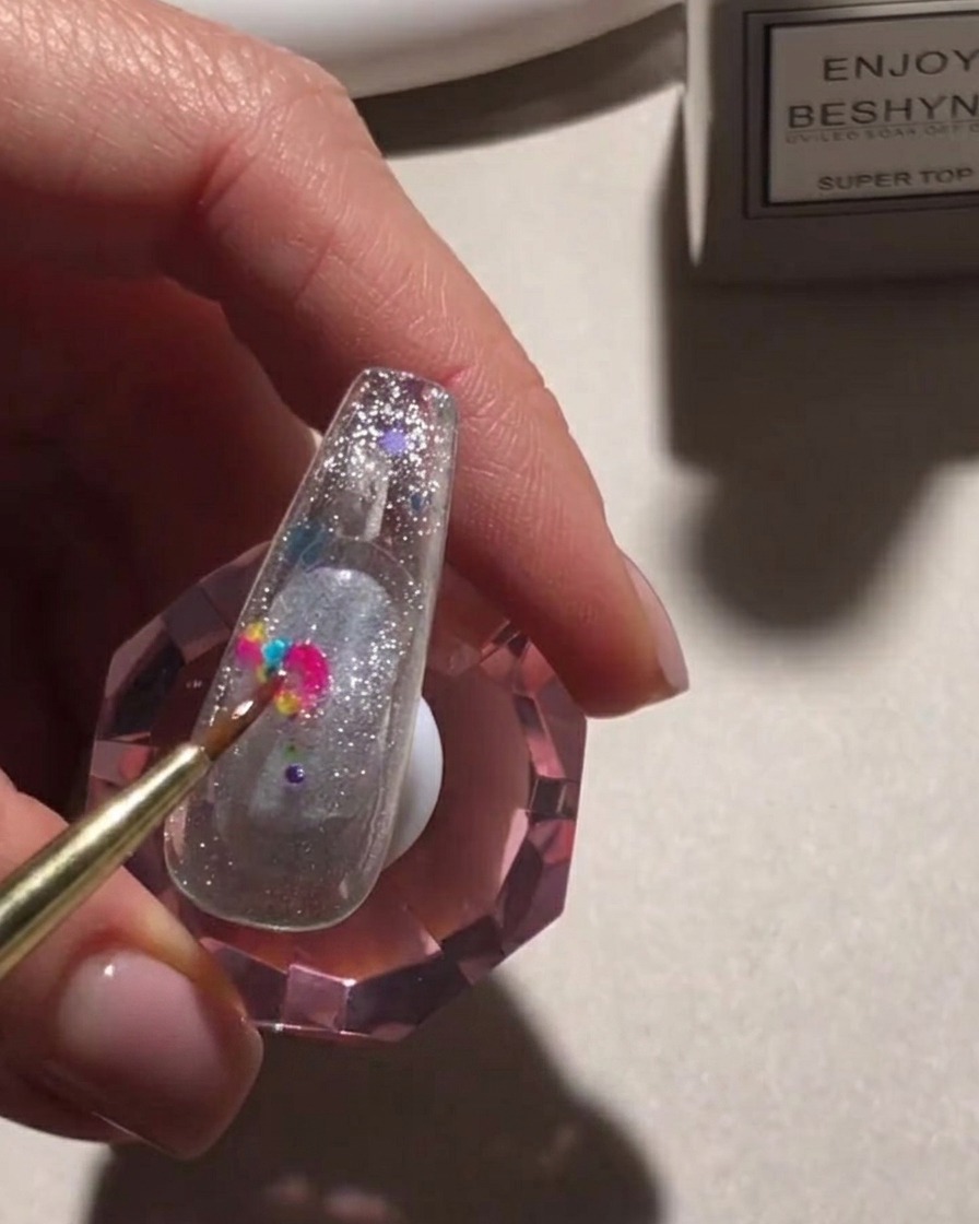 Ein Detailpinsel platziert bunte, runde Pailletten auf einer glitzernden Nageloberfläche.