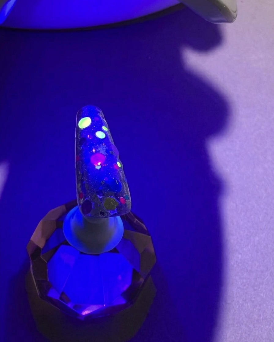 Ein Nagel mit bunten Pailletten unter dem violetten Licht einer UV-Lampe während des Aushärtevorgangs.