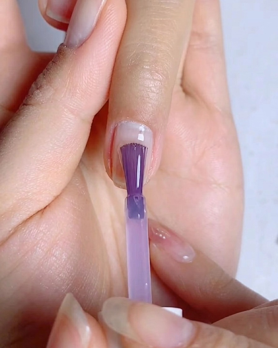 Ein Nagelpinsel verteilt eine dünne, gleichmäßige Schicht blau-violettes Gel auf der Nageloberfläche.