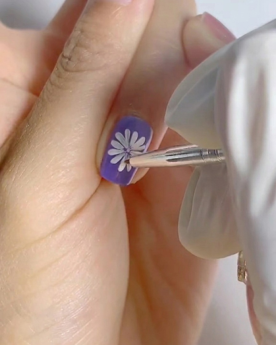 Ein Finger mit fertiggestelltem weißem Gänseblümchen-Design auf violettem Untergrund, der zur Aushärtung unter einer kleinen professionellen Nagellampe liegt.