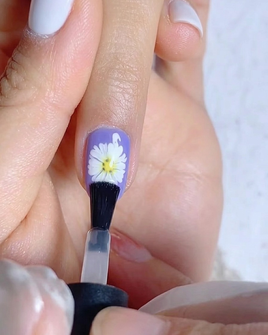 Ein Pinsel trägt eine Schicht matten Überlack auf einen violetten Nagel mit weißem Gänseblümchen-Design auf.