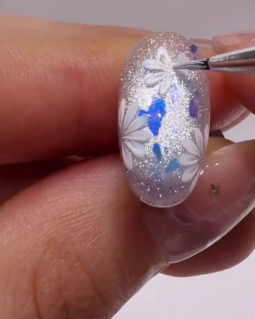 Ein feiner Liner-Pinsel malt präzise weiße Blütenblattformen auf einen glitzernden Nagel.