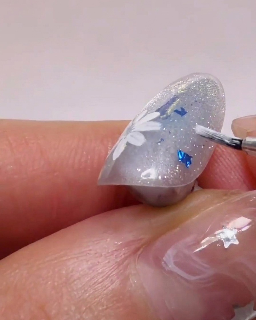 Ein Pinsel trägt weißes Gel auf, um die Form der Blütenblätter auf einem glitzernden Hintergrund zu ergänzen.
