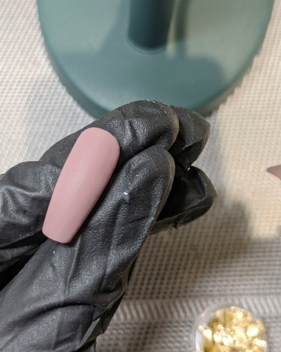 Ein Pinsel trägt eine Schicht mattes Top Coat über einen mit Blattgold verzierten Nagel auf.