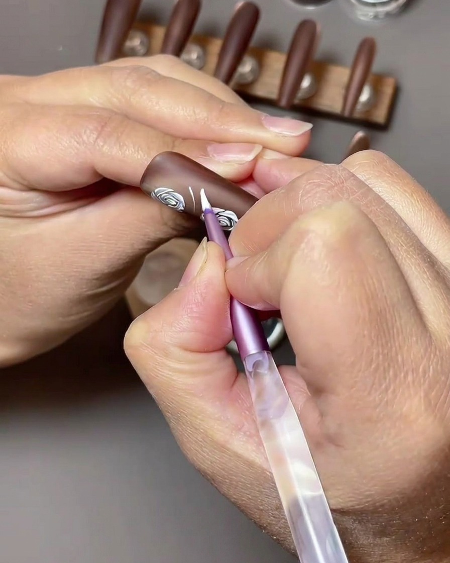 Nahaufnahme, wie weißes Nailart-Gel verwendet wird, um Blattformen neben bestehende weiße Rosen auf einen braunen Nagel zu malen.
