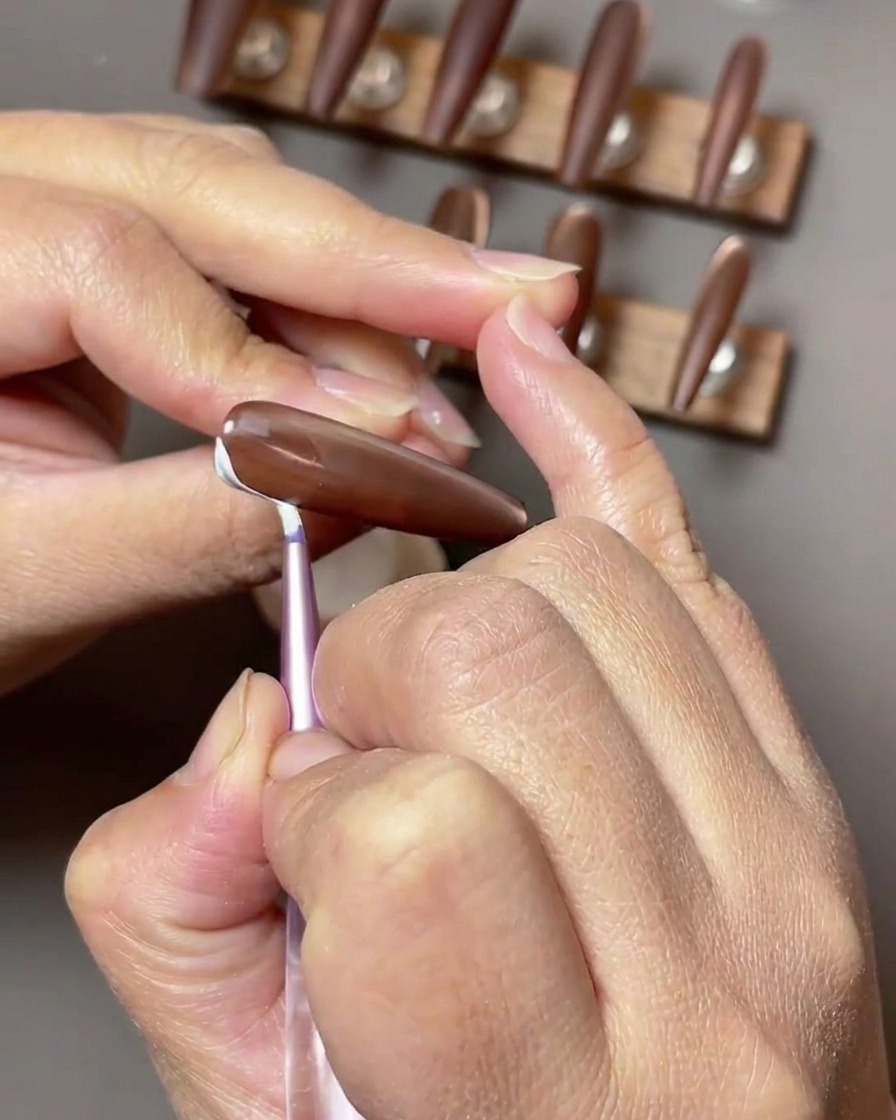 Nahaufnahme eines braunen Nagels, auf dem zarte weiße, bandartige Striche entlang der Nagelseite gemalt werden.
