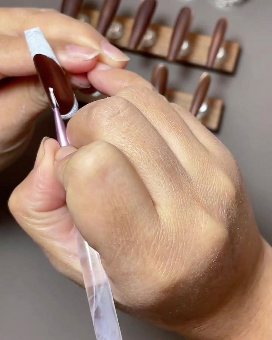 Nahaufnahme eines dünnen Nailart-Pinsels, der einen weißen vierzackigen Stern auf einen dunkelbraunen Nagel mit Reverse-French-Tip malt.