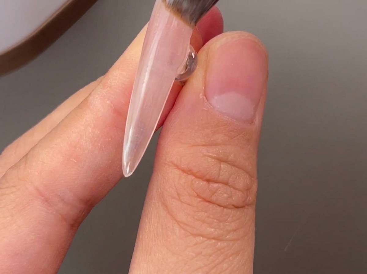 Ein Pinsel trägt eine klare Schicht Basisgel auf einen Naturnagel auf.