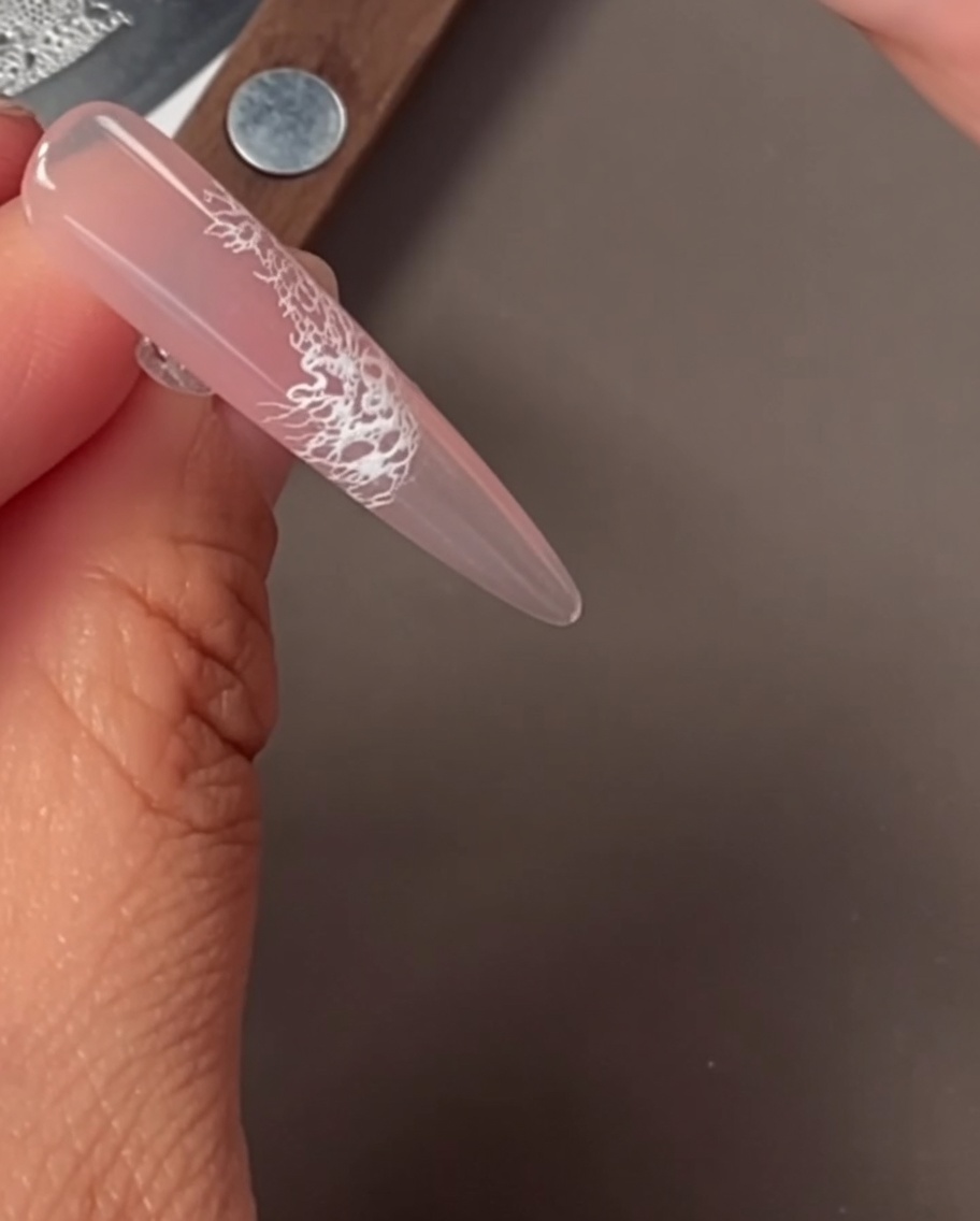 Nahaufnahme eines Nagels mit einem filigranen weißen Spitzenmuster, das per Stamping aufgetragen wurde.