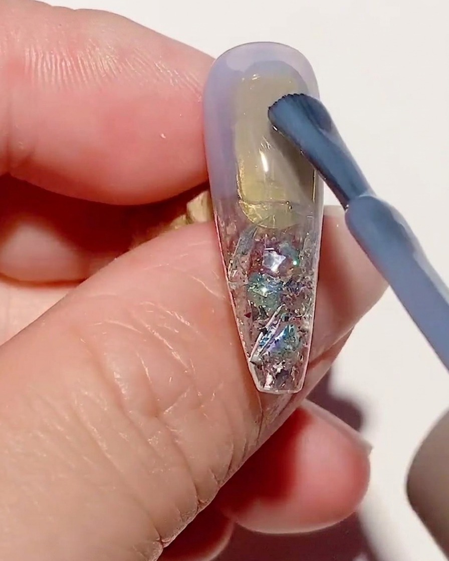 Ein Pinsel trägt eine dünne Schicht transparentes blaues Gel auf eine texturierte Nagelspitze auf.
