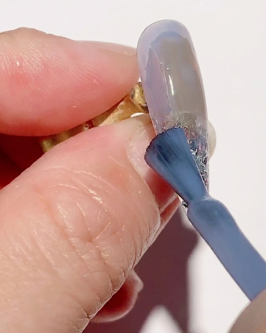 Ein Verlaufspinsel verblendet transparentes blaues Gel auf der Nageloberfläche für einen weichen Übergang.
