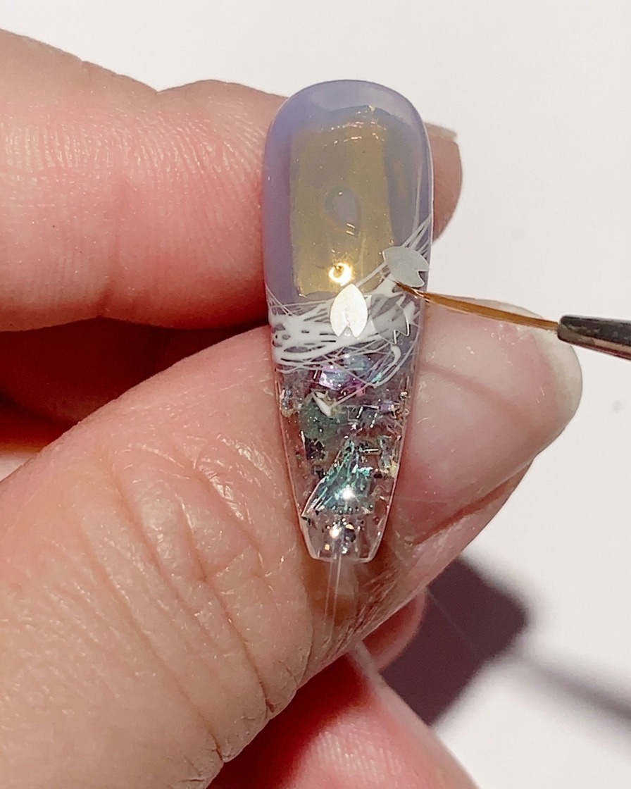 Eine Pinzette platziert kleine, filigrane kirschblütenförmige Pailletten auf der nassen Geloberfläche eines Nagels.