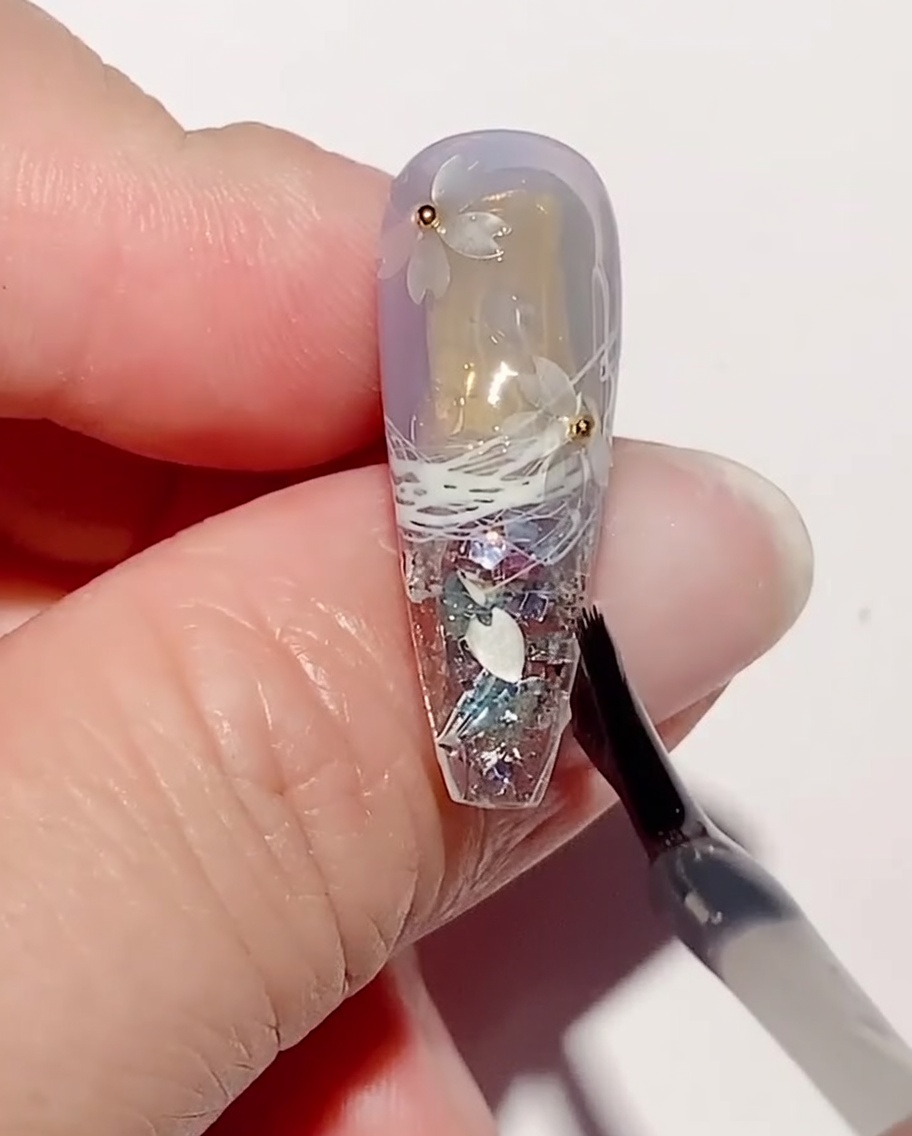 Ein Nailart-Pinsel trägt eine Schicht klares Verstärkungsgel über Kirschblüten-Pailletten und Stahlperlen auf einem Nagel auf.