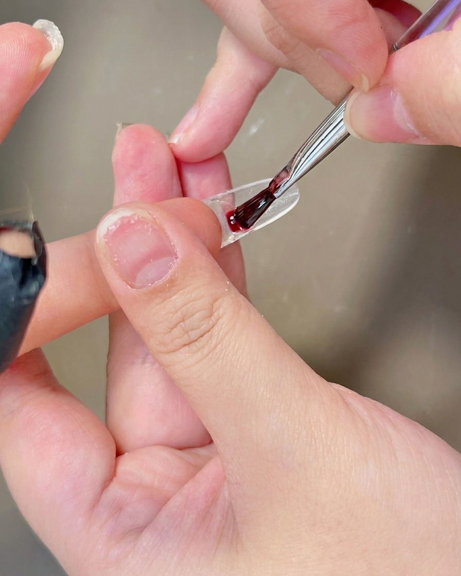 Ein Pinsel trägt eine Schicht transparentes, milchig-weißes Gel auf die Oberfläche eines klaren Nageltips auf.