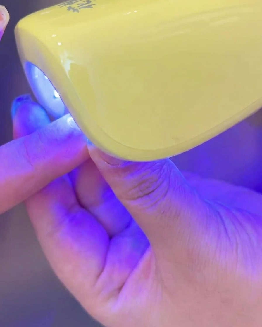 Eine Hand legt einen Nagel mit aufgetragenem milchigem Gel zum Aushärten unter eine kleine UV-/LED-Lampe.