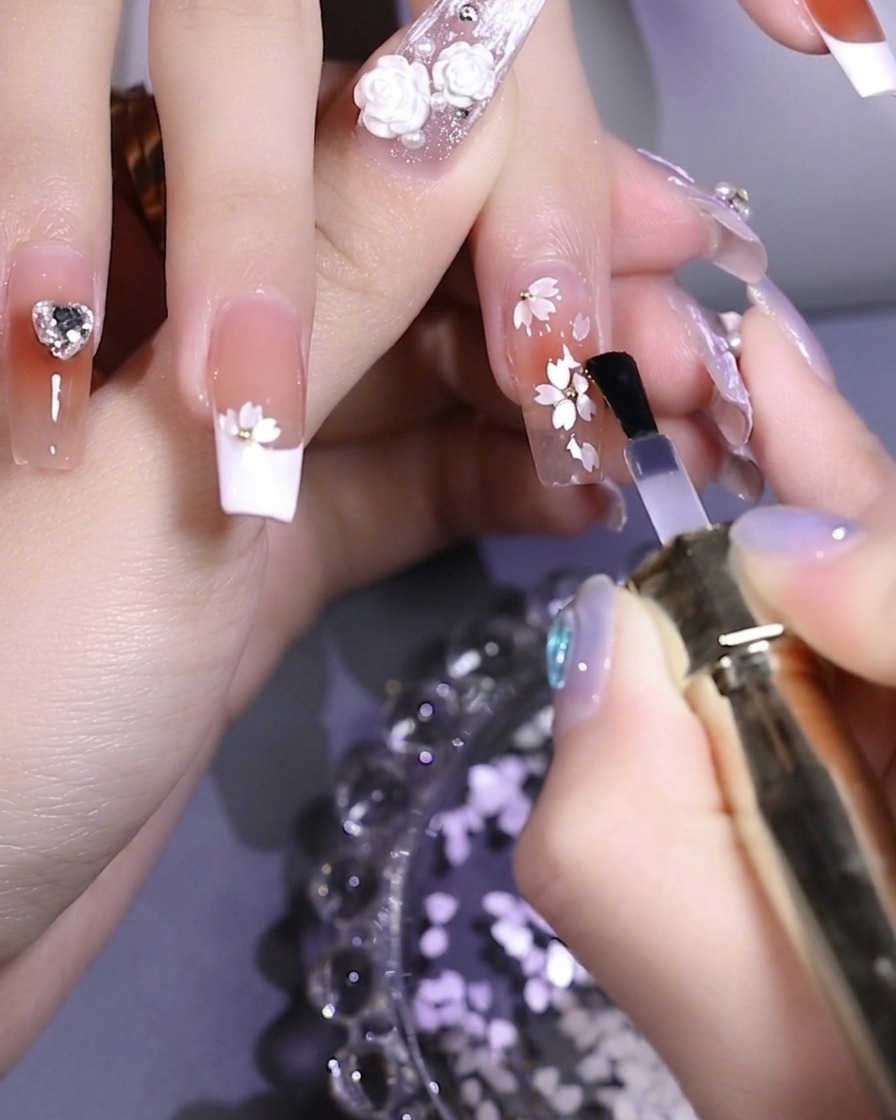 Ein Pinsel trägt klaren, glänzenden Top Coat auf einen mit weißen Pailletten und einer Metallperle verzierten Nagel auf.