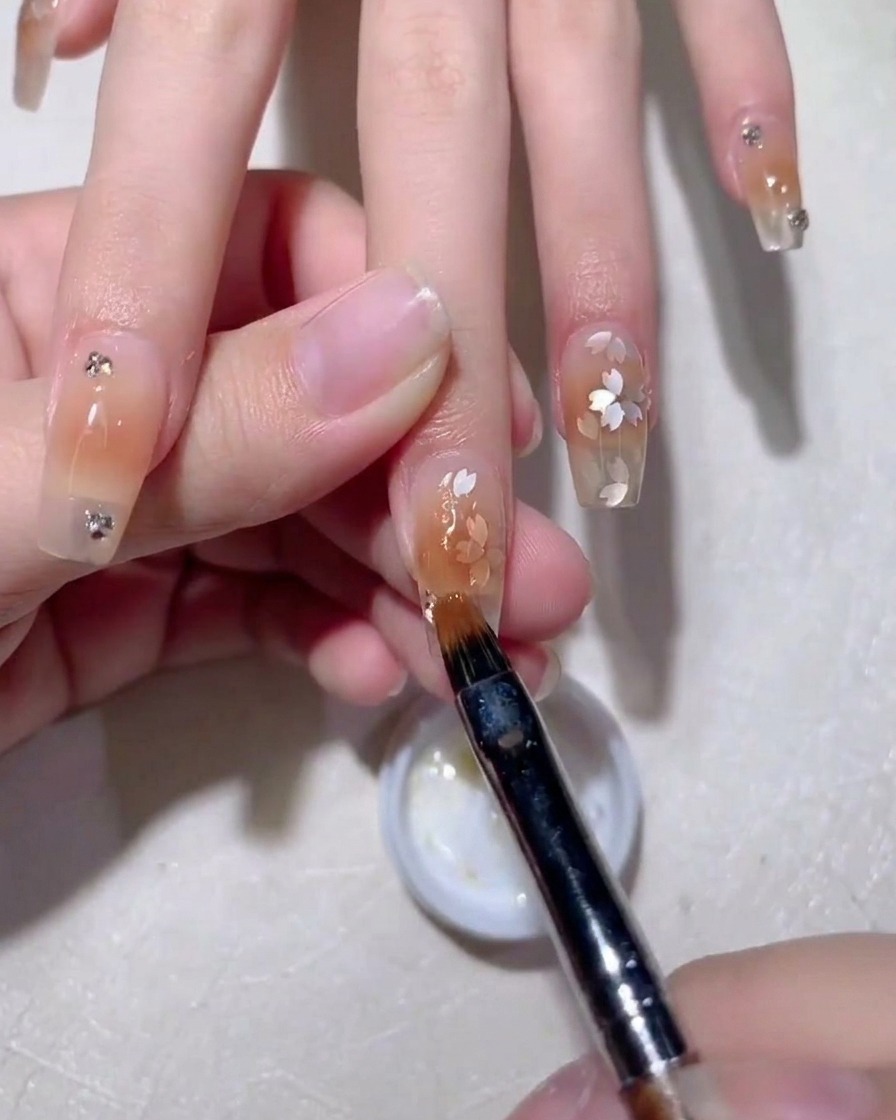Ein Nail-Art-Pinsel glättet klares Aufbaugel über Kirschblüten-Pailletten auf einem nudefarbenen Nagel.