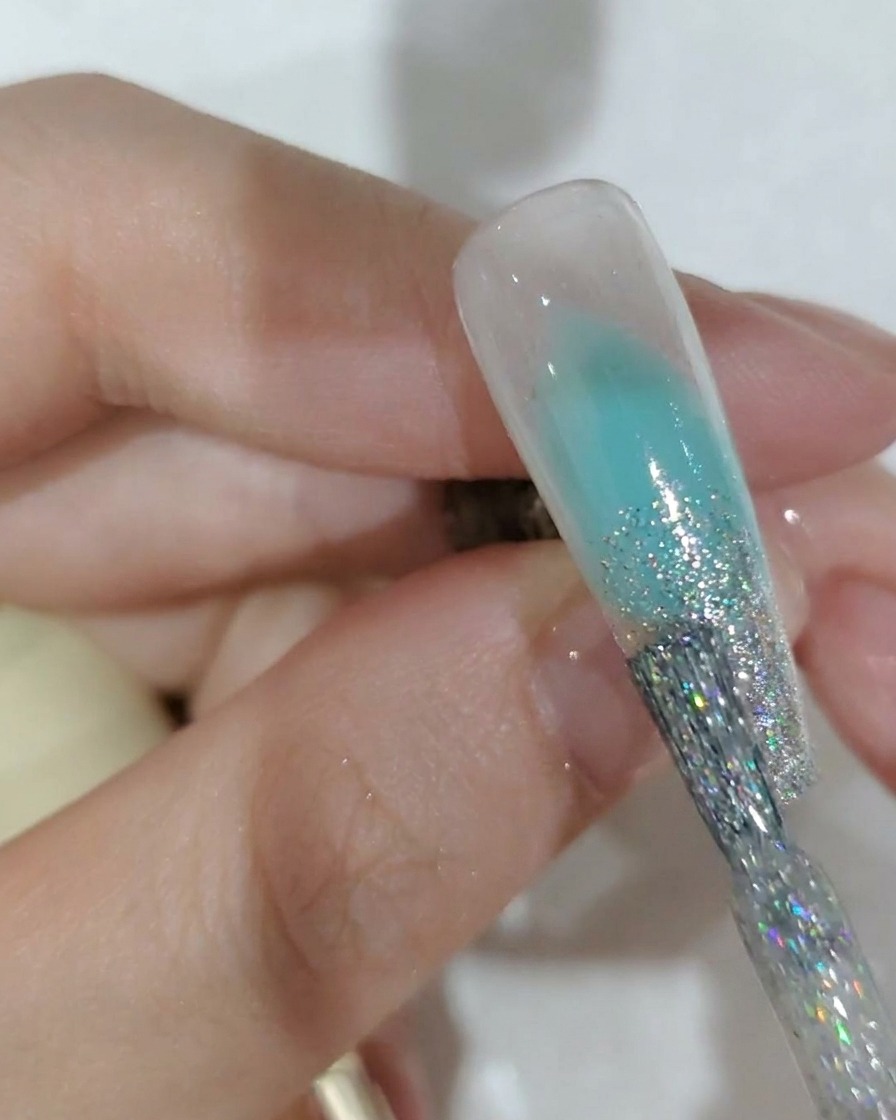 Nahaufnahme eines Nagelpinsels beim Auftragen von Silberglitzer-Gel auf die Nagelspitze.