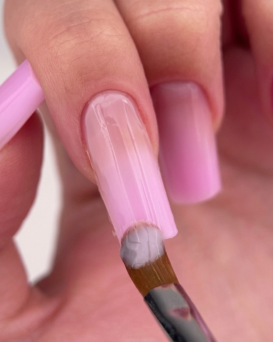 Ein Nagelpinsel trägt helles, milchiges rosa Gel nahe der Nagelhaut des Nagels auf.