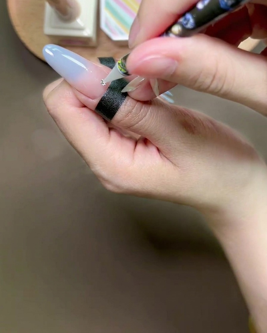 Ein Wachsstift platziert einen kleinen silbernen Strassstein auf einem Klebetropfen auf einem manikürten Nagel.
