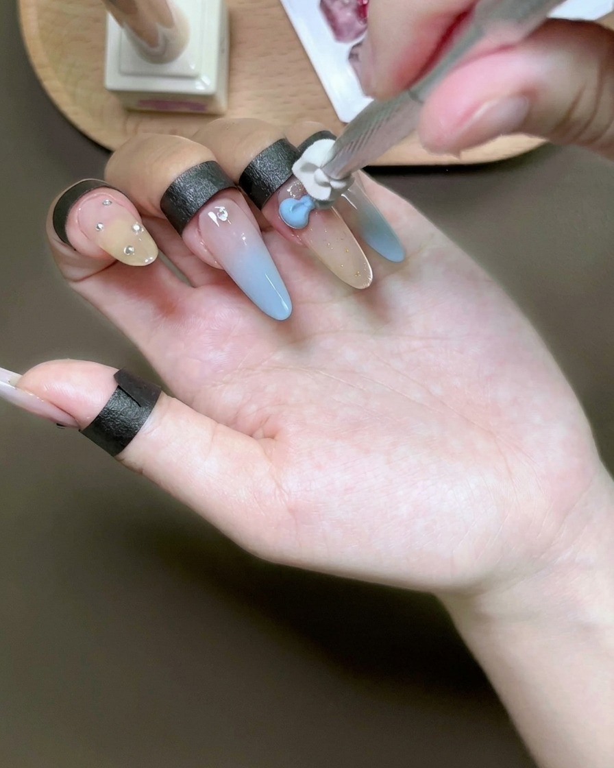 Ein Schwämmchen tupft hellblaues Gel über eine weiße 3D-Schleife auf einem Fingernagel.