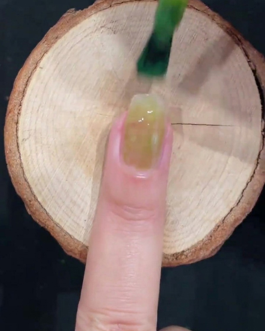Ein Gel-Pinsel trägt halbtransparentes, eisgrünes Gel auf die Mitte eines Naturnagels auf.