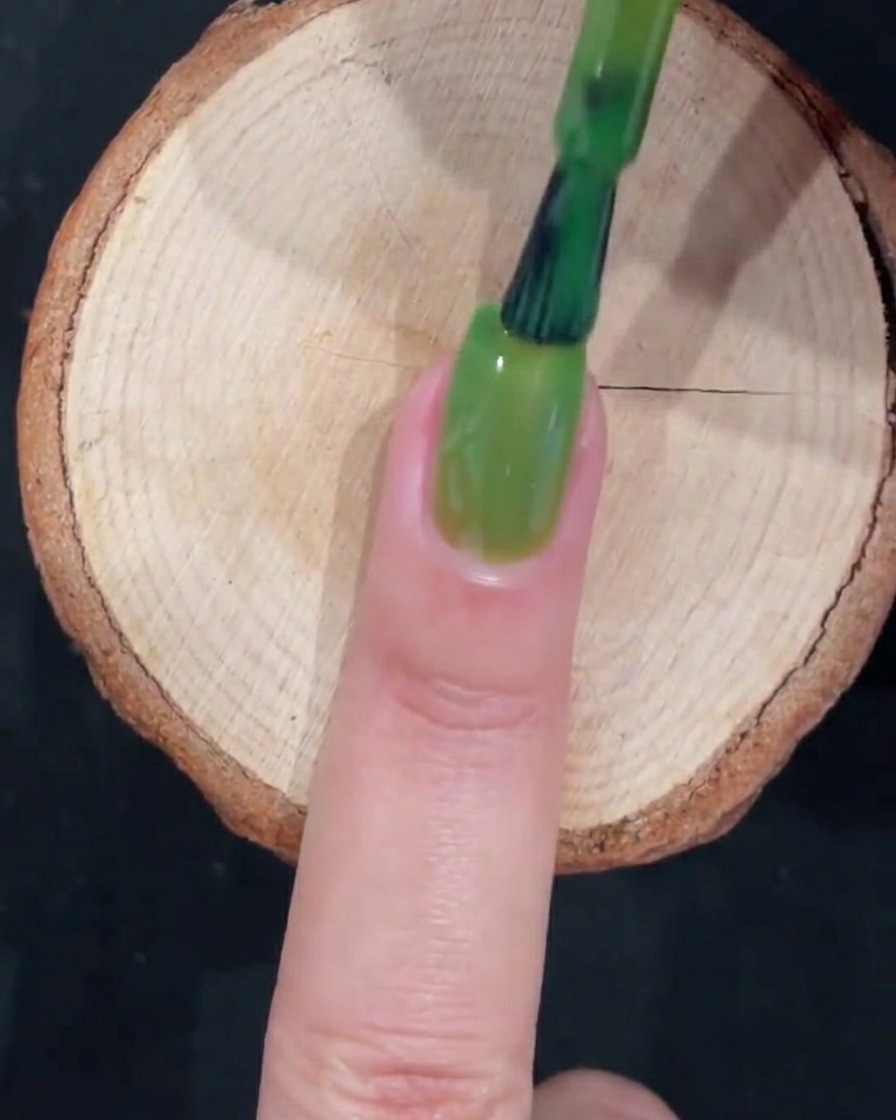 Ein Gel-Pinsel zieht eine glatte Schicht transparentes grünes Gel bis zur Nagelspitze.