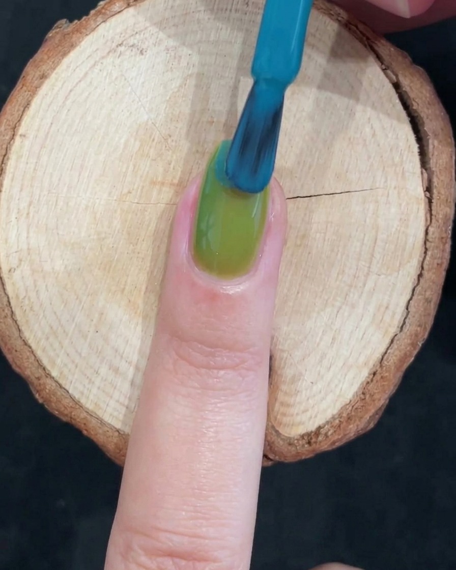 Ein Nagelpinsel trägt kräftiges blaues Gel auf die obere Hälfte des grünen Nagels auf.