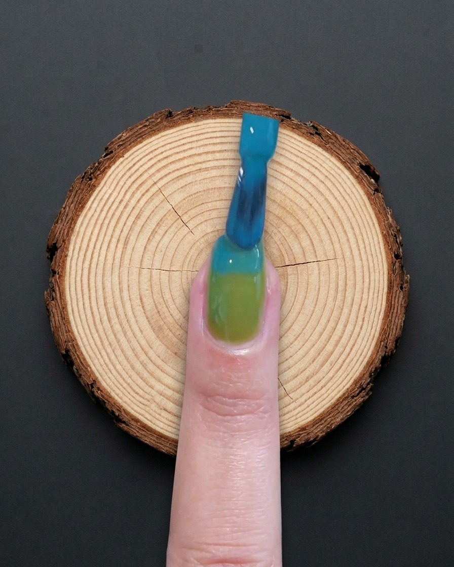 Ein Pinsel verteilt blaues Gel auf der oberen Hälfte eines grünen Nagels.