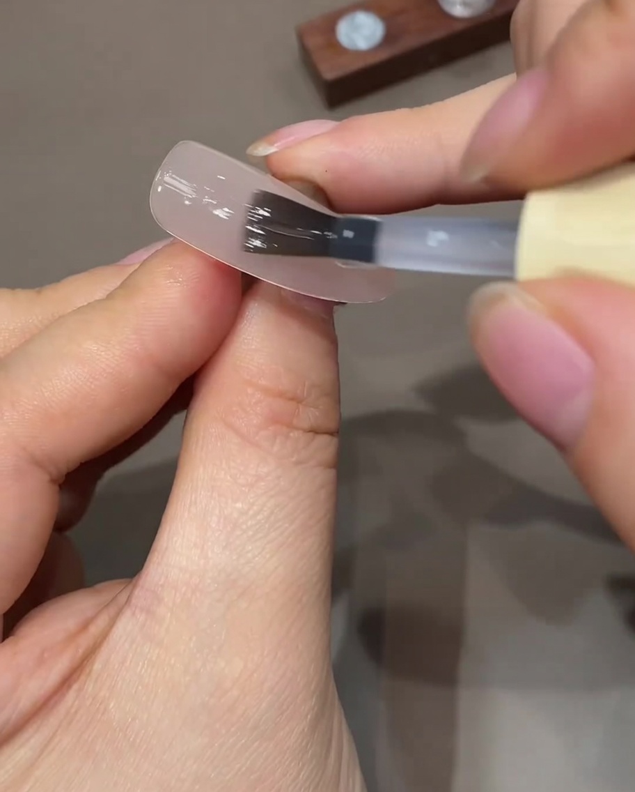 Ein flacher Gel-Pinsel trägt eine dünne Schicht Top Coat auf einen vorbereiteten Nude-Nagel auf.