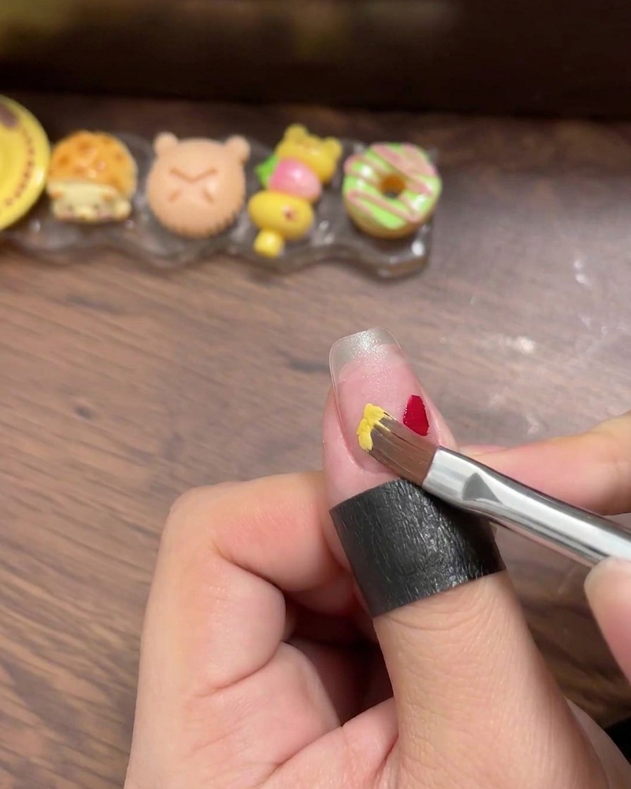 Nahaufnahme, wie mit einem Liner-Pinsel kleine, deutliche Quadrate aus rotem, gelbem, blauem und mintfarbenem Gel-Nagellack auf eine Nagelspitze aufgetragen werden.