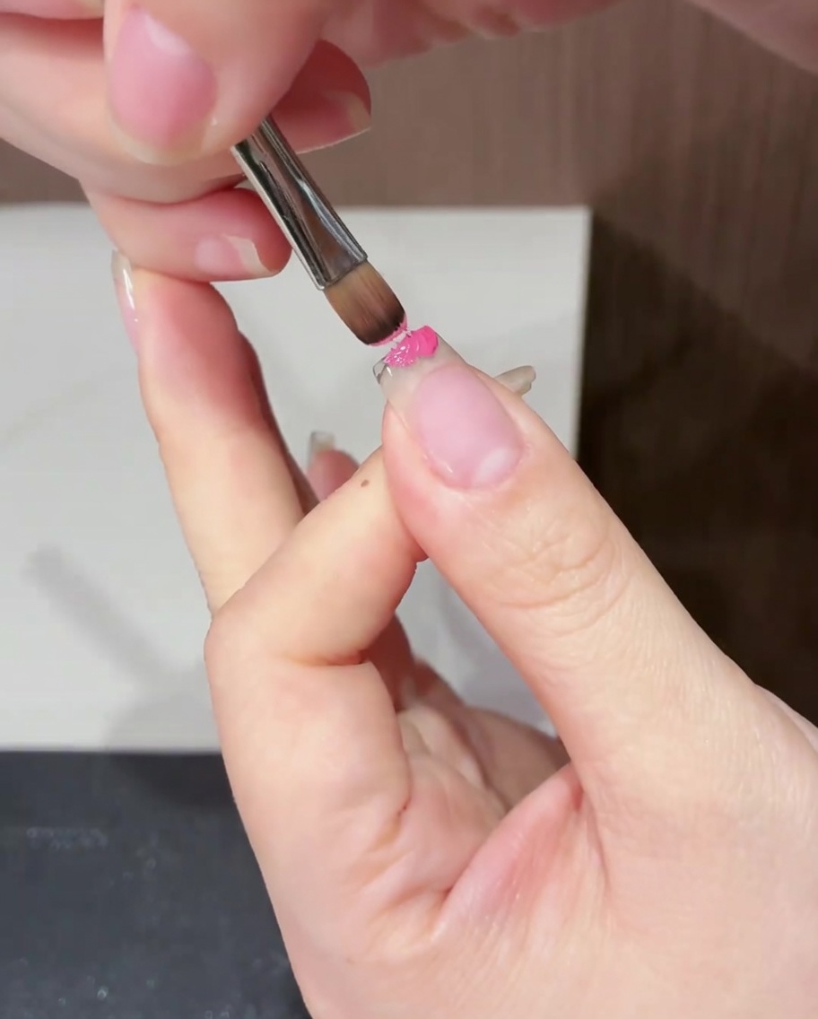 Nahaufnahme eines flachen Pinsels, der magentafarbenen Gel-Lack gezielt auf den freien Rand eines Naturnagels aufträgt.