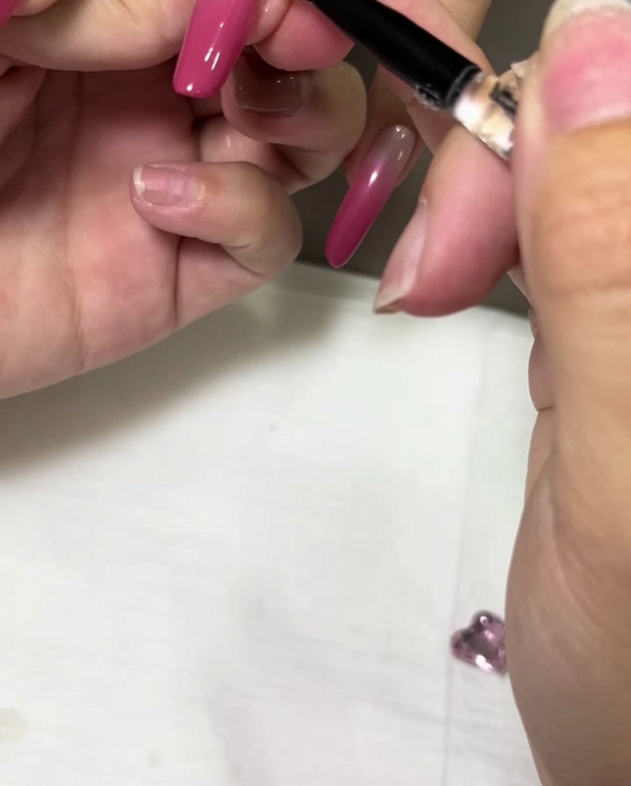 Ein feiner Detailpinsel trägt einen Tropfen klaren Gel-Kleber auf einen rosa Ombré-Nagel auf.