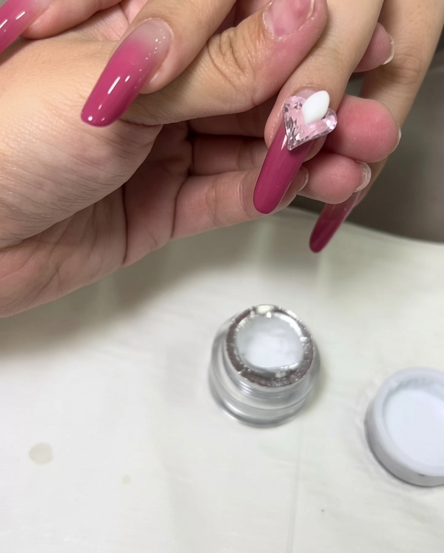Eine kleine Kugel weißes 3D-Modelliergel wird neben einem rosa Herzkristall auf den Nagel gesetzt.